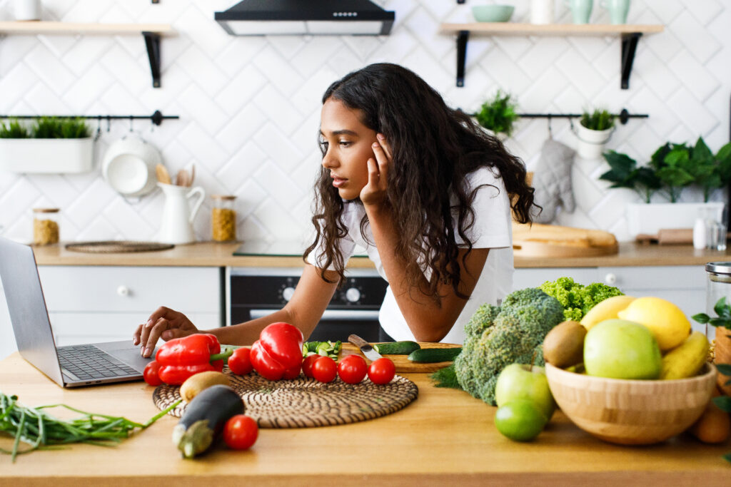 Thoughtful beautiful mulatto girl is looking on the laptop screen  on the modern kitchen on the table full of vegetables and fruits, dressed in white t-shirt